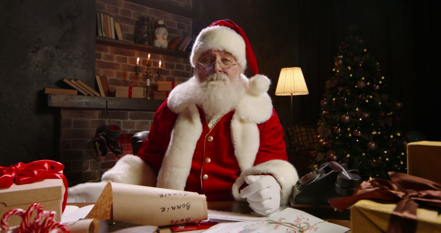 Santa Claus holding New Year present close-up. Talking on video call, using virtual conference, congratulating online, showing gift. Grey-haired Saint Nicholas working at night on Christmas Eve. 