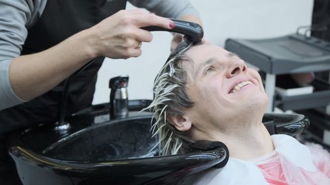 A handsome young man with a smile is resting in a hairdressing salon. After dyeing the hair white, the hairdresser thoroughly washes the young man's head with water. Young man happy with the result of