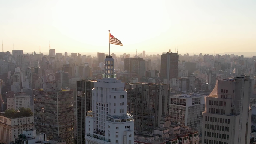 Altino Arantes building. Sao Paulo flag waving at rooftop. Sunset sky at downtown Sao Paulo. Downtown sunset cityscape of São Paulo, Brazil. Skyline downtown city. Santander Lighthouse building flag. Royalty-Free Stock Footage #1081261370