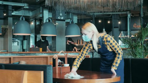 Female waitress in a face mask is doing a clean-up in the restaurant. Small business during covid-19, coronavirus pandemic.
