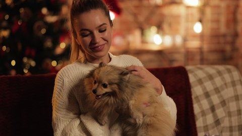 Cute Happy Young Woman is playing with her lovely little Dog, Puppy. Young Female is celebrating Christmas, New Year at Home with her Dog. Cozy Festive Evening. Happy Holidays. Christmas Spirit.
