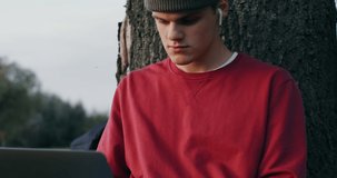 A young man of European appearance, in a knitted hat, uses a laptop, sitting on the ground under a tree in the open air.
