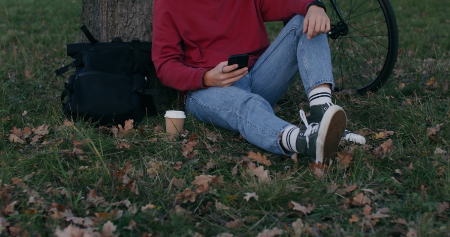 A young man of European appearance uses a phone while sitting on the ground under a tree, then gets up, collecting his things and leaves, taking the bike in his hands