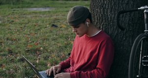 A young man of European appearance in a knitted hat and wireless headphones uses a laptop sitting on the ground under a tree. The bike stands next to the guy