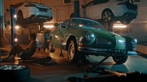 Caucasian female mechanic repairing a vintage old car in a workshop, working under car bottom. Shot with 2x anamorphic lens