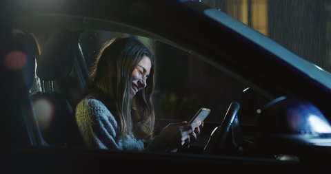 Cinematic shot of young happy woman using smartphone for send messages and navigate in internet on driver seat alone while waiting to stop raining before departure to her destination at night.