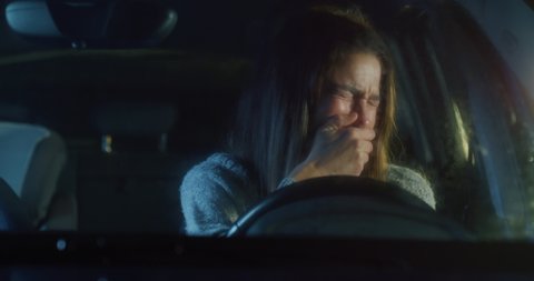 Cinematic shot of young desperate worried upset sad suffering from severe depression heart broken woman is crying with tears coming from eyes and falling on cheeks alone on driver's seat of her car.