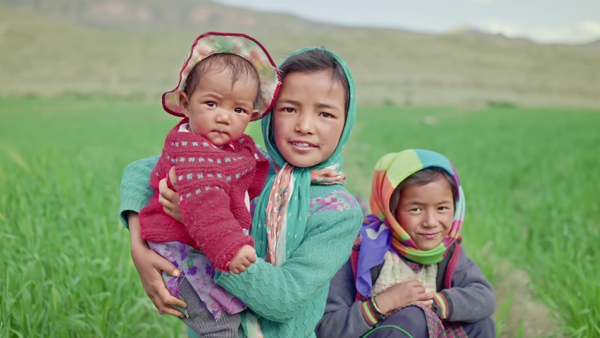 Shot of a young little rural girl holding a cute infant female baby accompanied by a little sister next to an agricultural field in the mountainous region looking at the camera and smiling.  Royalty-Free Stock Footage #1081290065