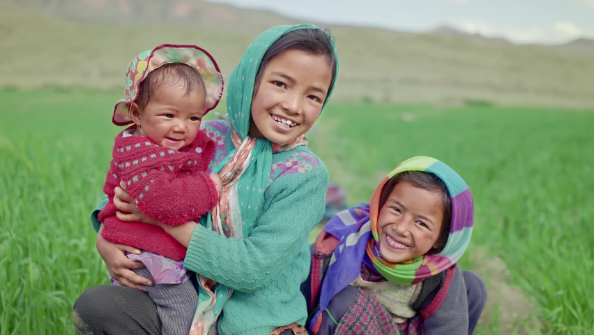 Shot of a young little rural girl holding a cute infant female baby accompanied by a little sister next to an agricultural field in the mountainous region looking at the camera and smiling.  Royalty-Free Stock Footage #1081290065