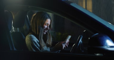 Cinematic shot of young happy woman using smartphone for send messages and navigate in internet on driver seat alone while waiting to stop raining before departure to her destination at night.