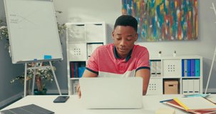 A young boy of African American beauty sits at desk in an office room with a laptop, smiles broadly and waves hand, carries on a conversation via internet, video, has remote teaching