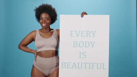Medium studio portrait of confident plus size African-American woman in underwear posing for camera holding Every Body Is Beautiful poster standing on blue background