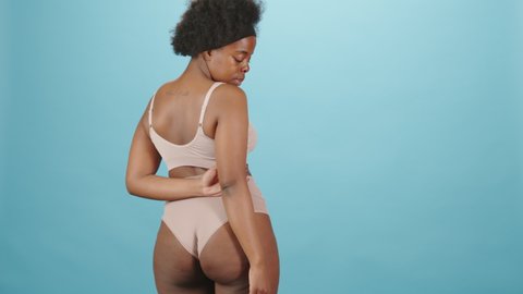 Rear-view medium portrait of young plus size African-American woman in underwear posing for camera showing her natural healthy body standing on isolated blue background