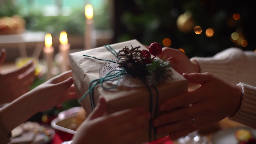 Close-up hands of mother giving festive box with Christmas present to son sitting at dinner feast table during holiday family party, decorating xmas tree and celebration lights, selective focus. Royalty-Free Stock Footage #1081300730