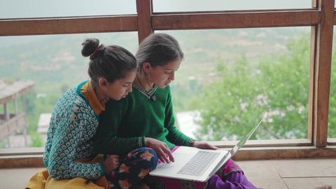 Two young village or rural school girls sitting indoors and using the laptop with the internet to study in an interior house with the green mountains in the back. learning and female education concept