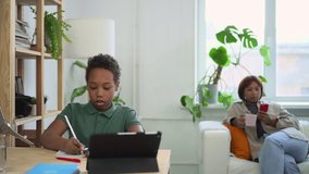 Boy learning in front of tablet screen, mother sitting on couch with phone in hand at home spbd. African American schoolboy writes in notebook and looks with smile, young woman uses smartphone and