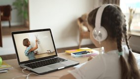 Online education at home. Little girl pupil with modern headphones listens to the teacher at English video lesson via notebook at table backside view