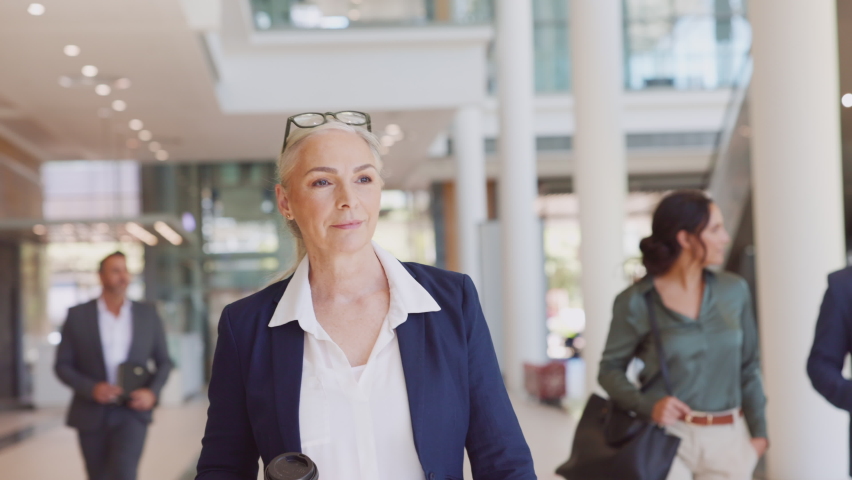 Successful mature businesswoman in modern business centre walking and looking proudly at camera. Smiling senior woman walk with positive expression and holding coffe cup with people in background. Royalty-Free Stock Footage #1081307804