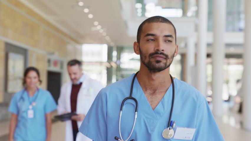 Portrait of smiling middle eastern nurse in uniform walking at hospital hallway. Young doctor walking in hospital corridor with health care team in background. Successful Indian surgeon working. | Shutterstock HD Video #1081307822