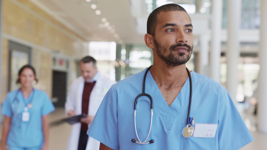 Portrait of smiling middle eastern nurse in uniform walking at hospital hallway. Young doctor walking in hospital corridor with health care team in background. Successful Indian surgeon working. | Shutterstock HD Video #1081307822