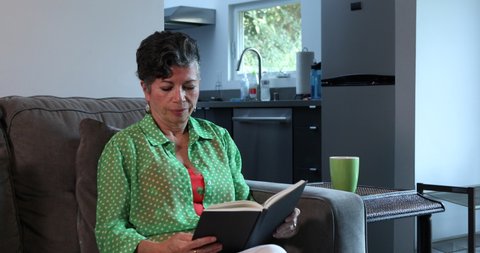Hispanic woman reads book at home and sips tea.