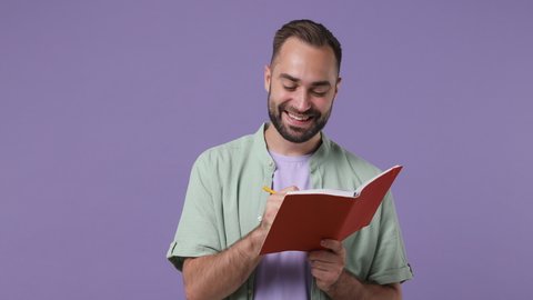 Smiling dreamful pensive thoughtful wistful minded young bearded man 20s years old wears mint shirt hold write fill in brown notebook diary isolated on plain light purple background studio portrait
