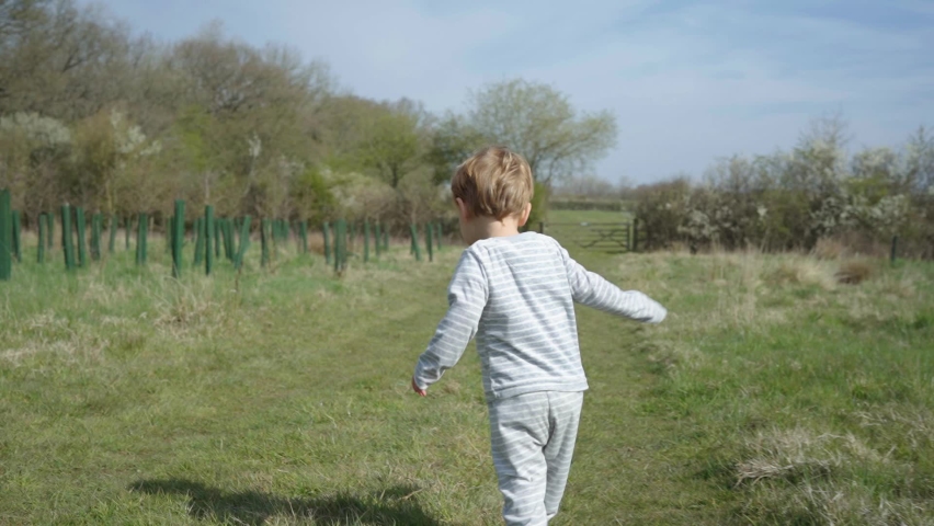 Toddler boy runs through field along young trees on sunny day, slow motion