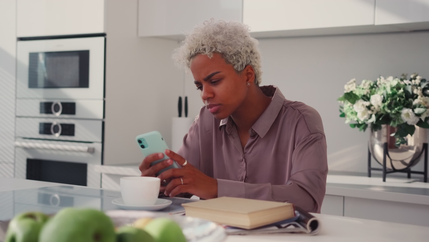 Worried Latina woman having problems with mobile phone sitting in kitchen, using wrong working apps getting annoyed with spam reading message with bad news feeling stress, angry about cell breakdown. Royalty-Free Stock Footage #1081333547