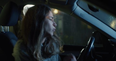 Cinematic authentic shot of young happy woman is having fun to dance and sing crazy on driver seat alone while waiting to stop raining before departure to her destination at night.