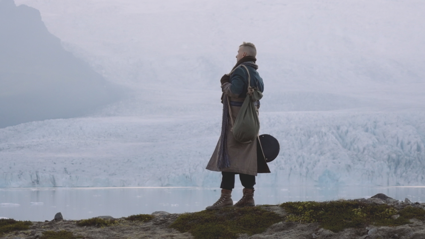 Lockdown Wide Slow Motion Shot Of Man With Guitar Case And Greatcoat Standing By Lake With Glacier, Iceland