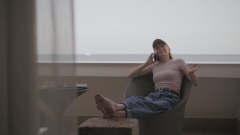 A woman in a baseball cap is talking on the phone sitting on the balcony of a hotel room with a sea view.