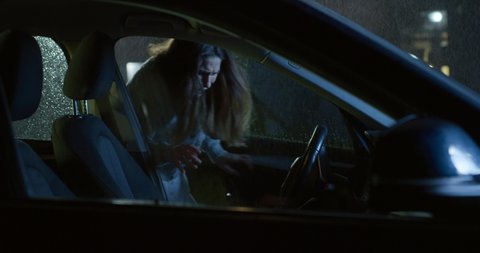 Cinematic shot of young woman covering her head from rain and running to enter in car on driver seat before departure to her destination at night.