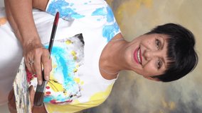 A mature female artist poses against the background of her painting in the studio. Vertical video for smartphone screen, for target social media platforms and web browsers on mobile devices.