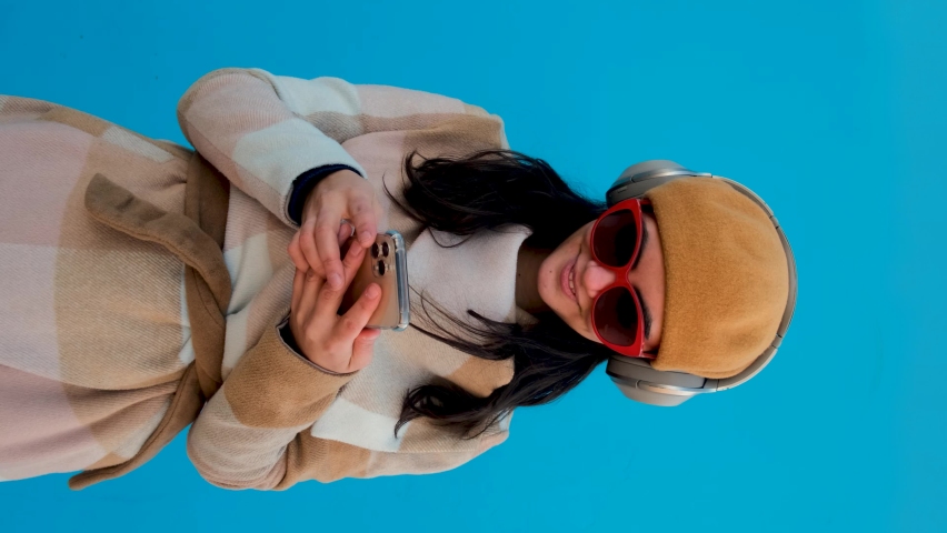 Positive female with black hair in sunglasses and hat listening to music in headphones while surfing cellphone on blue background