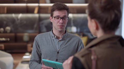 Portrait of confident young male seller in eyeglasses with tablet standing in furniture shop talking to client. Expert Caucasian salesman consulting customer in luxurious store showroom indoors