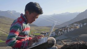 A young East Asian rural boy sitting outdoors in the morning sunlight engrossed studying in a laptop with a beautiful landscape view of a mountainous region with a tiny village on its foothills.