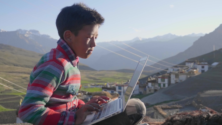 A young East Asian rural boy sitting outdoors in the morning sunlight engrossed studying in a laptop with a beautiful landscape view of a mountainous region with a tiny village on its foothills. | Shutterstock HD Video #1081355354