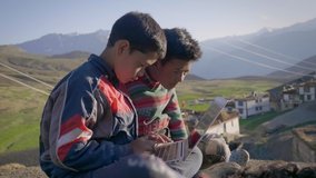 Two young East Asian rural kids sitting together outdoors in the morning engrossed in learning on a laptop with a beautiful landscape view of a mountainous region with a tiny village on its foothills 