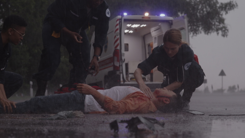 Asian man lying injured on a road under strong rain and getting professional medical care from team of ems paramedics. Multicultural doctors in hurry. First aid for victim.