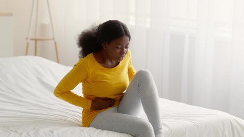 Period Pain. Sick Black Woman Suffering From Abdominal Ache At Home, Young African American Lady Sitting On Bed And Rubbing Belly, Having Menstrual Cramps Or Digestive Problems, Slow Motion Footage