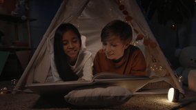 Teenagers in decorative makeshift hut reading book and smiling at home in evening. Positive boy and girl lying on floor while spending free time. Concept of leisure and carelessness - Powered by Shutterstock - Get 15% off with code: PIKWIZARD15