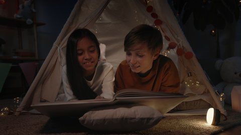 Teenagers in decorative makeshift hut reading book and smiling at home in evening. Positive boy and girl lying on floor while spending free time. Concept of leisure and carelessness