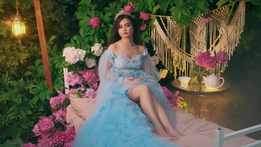 Fairy fantasy woman woke up sitting on bed. girl beauty princess in carnival blue vintage dress. Brunette queen long flowing hair, crown on head. pink flowers, green peony leaves, fog smoke in garden