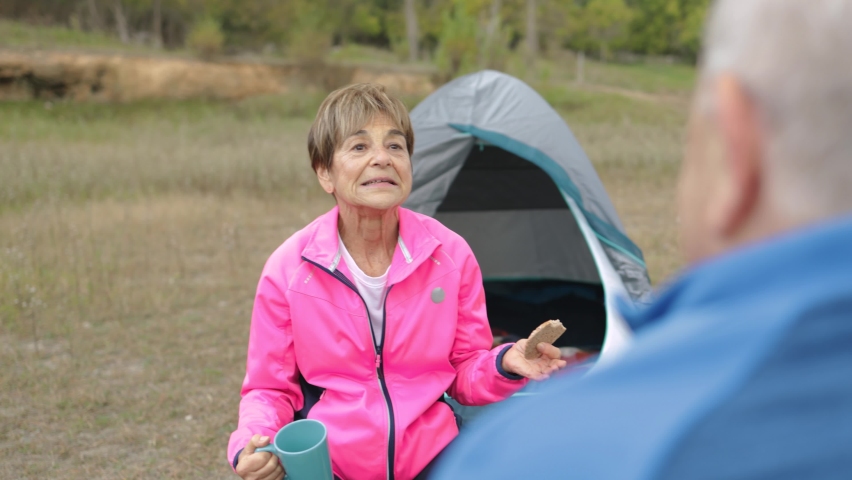 Happy senior couple having fun eating and drinking while camping outdoor in the woods - Elderly travel vacation
