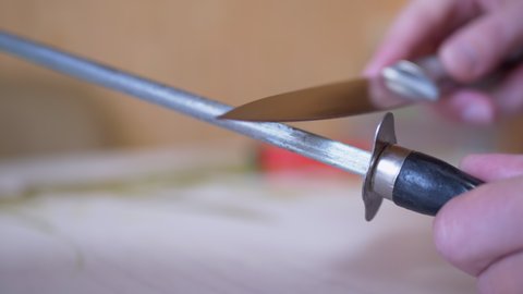 Female Hands Sharpens a Knife on Old, Dirty, Scratched a Steel Rod in Kitchen. Dull knife blade. Preparation of the working tool for cutting. Blurred background. Daylight illumination. 4K. Close up.