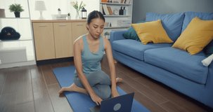 Young attractive Asia woman in sportswear site on mat in the floor watching fitness online video on laptop practice yoga for beginner in living room at house. Home quarantine and fitness exercise.