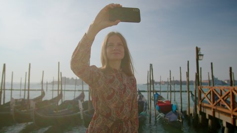 Beautiful Woman Takes Selfie Photos by Phone with Gondolas in Venice Grand Canal, Italy, Europe. Young Lady Tourist Travels alone at Pandemic. Blogging and Landmark Sightseeing Concept. 4K orbit shot