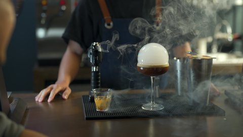 Asian man barista shaking iced black coffee in shaker and pouring in cocktail glass on bar counter at coffee shop. Male coffee shop owner serving cold drink coffee to customer. Small business concept.