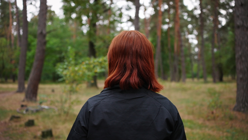 Unrecognizable redhaired sporty woman putting on protective helmet before extreme bicycle ride in autumn forest, safe workout concept, slow motion, back view