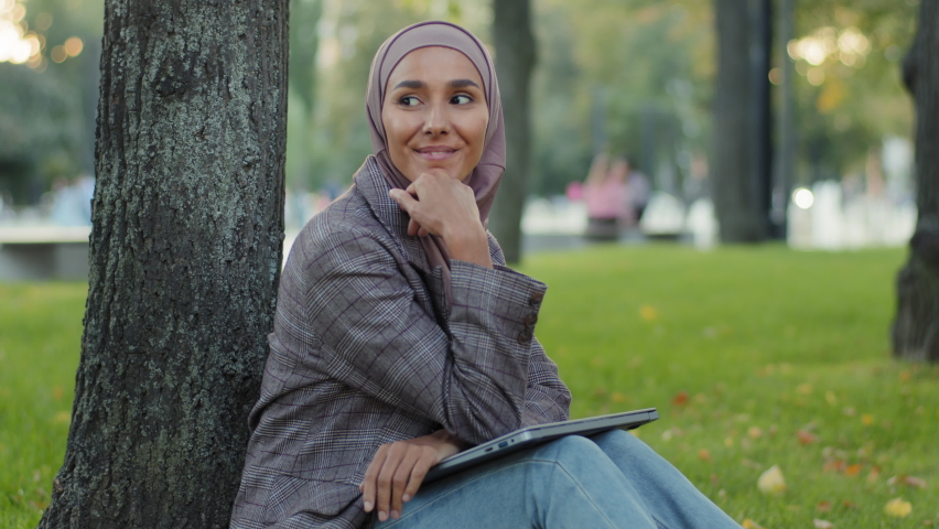 Smiling dreamy creative pensive girl islamic muslim business woman female writer journalist in hijab sitting on green grass lawn in park coming up with new novel typing on laptop article inspiration Royalty-Free Stock Footage #1081379453